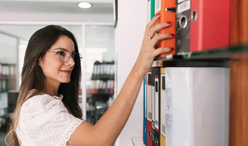 Das Bild zeigt eine Frau im Büro, die einen Aktenordner aus dem Schrank holt.