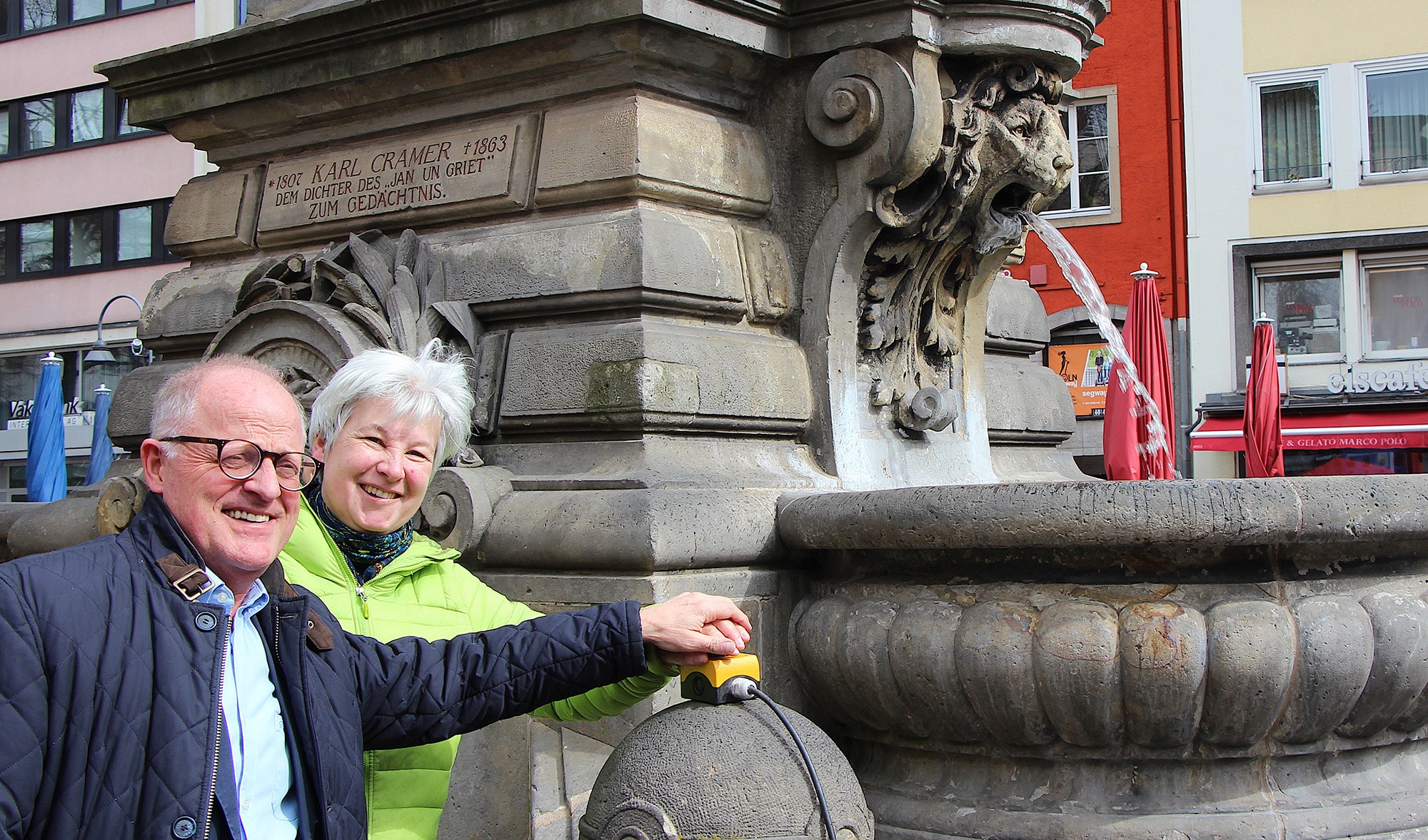 Das Foto zeigt zwei Personen, die das Wasser des Jan-van-Werth-Brunnen auf dem Alter Markt mit einem Knopfdruck sprudeln lassen.