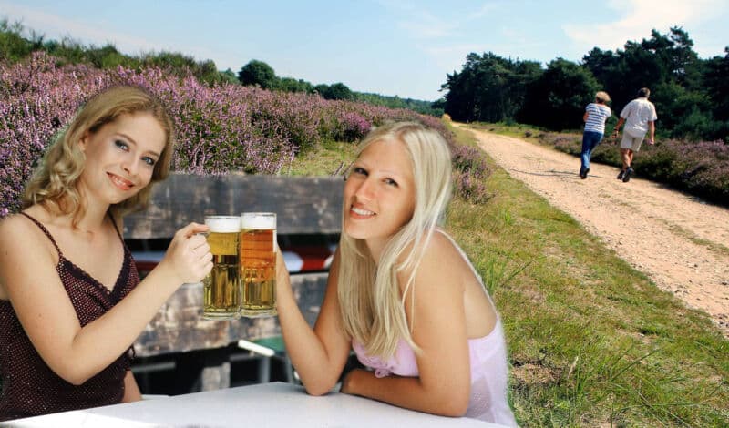 Zwei junge Frauen stoßen mit einem Bier an. Im Hintergrund sieht man Wanderer in der Wahner Heide.