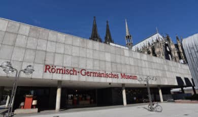 Das Foto zeigt die Außenfassade des Römisch-Germanischen Museum vor der Kulisse des Kölner Doms.