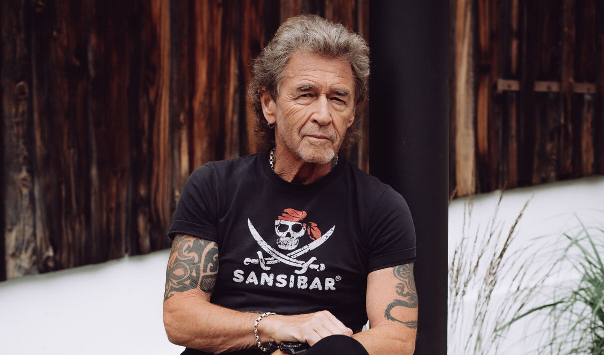 Peter Maffay sitzt im T-Shirt vor einer Holzwand