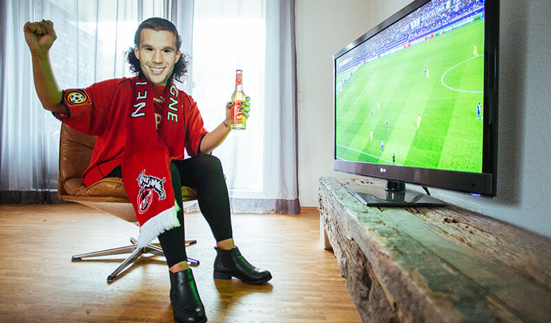 Person sitzt verkleidet als Lukas Podolski auf einem Stuhl vor einem Fernseher.