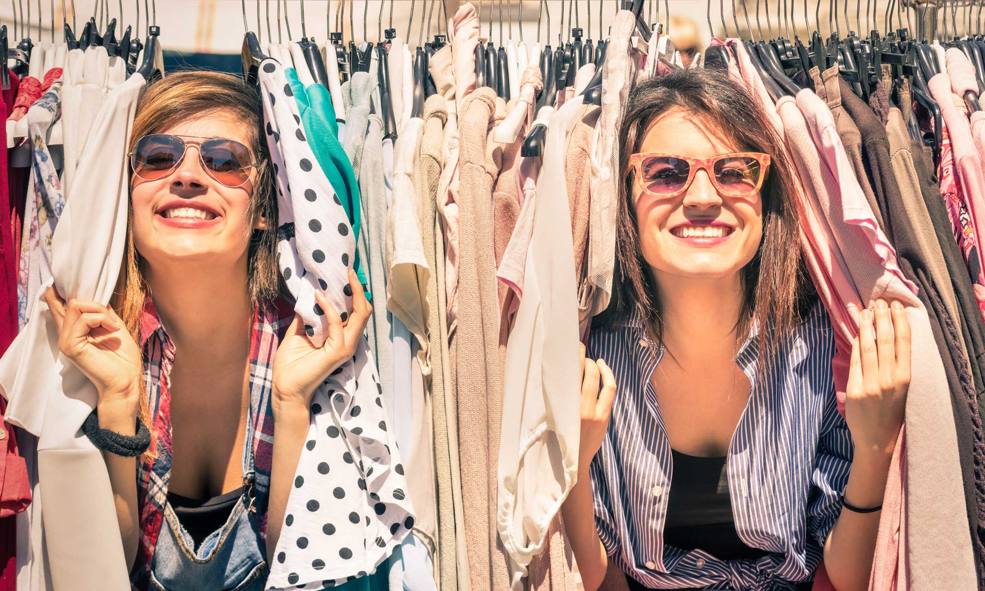 Zwei junge Frauen gucken zwischen Kleiderständern mit Mode hervor