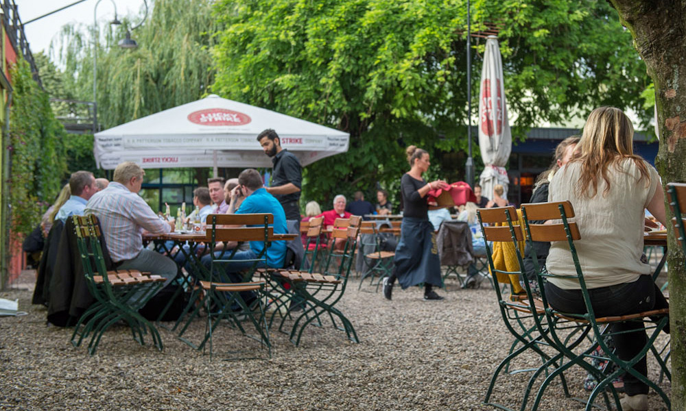 Besucher sitzen im Biergarten Herbrand's, eine Kellnerin läuft unter Bäumen.