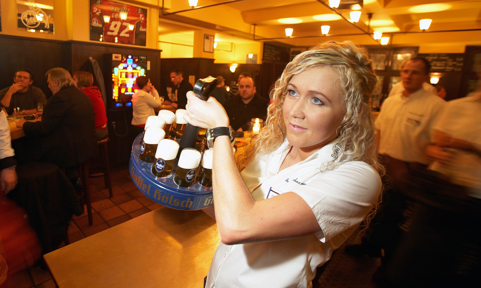 Eine Kellnerin trägt einen Kranz mit Kölsch durch das Brauhaus ohne Namen