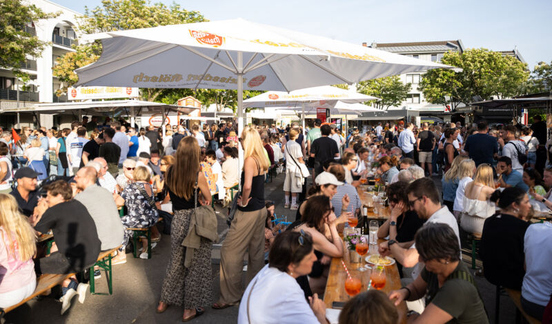 Beim Feierabendmarkt auf dem Hermeskeiler Platz in Köln-Sülz sitzen Menschen auf Holzbänken an Tischen