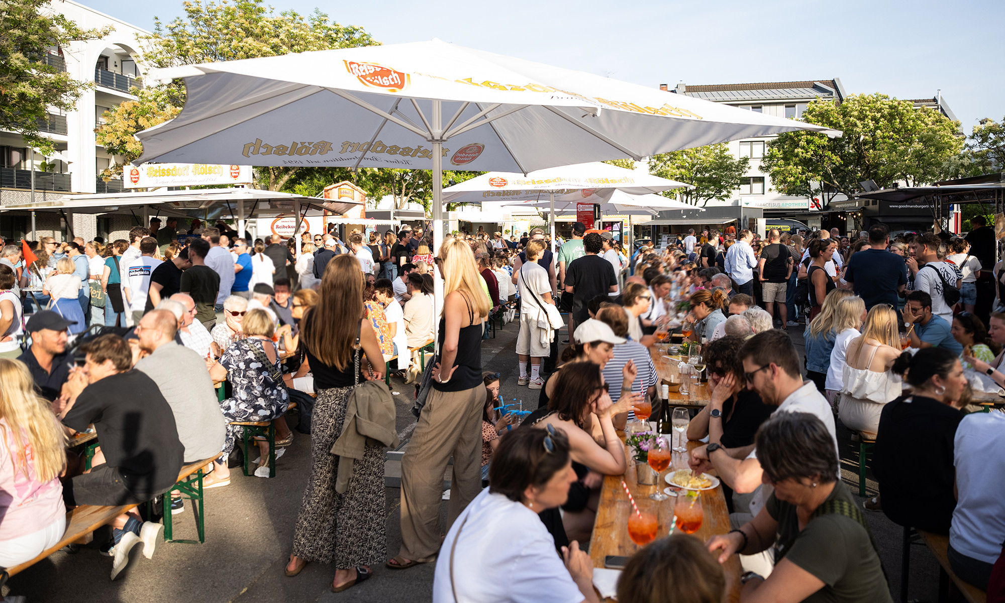Beim Feierabendmarkt auf dem Hermeskeiler Platz in Köln-Sülz sitzen Menschen auf Holzbänken an Tischen