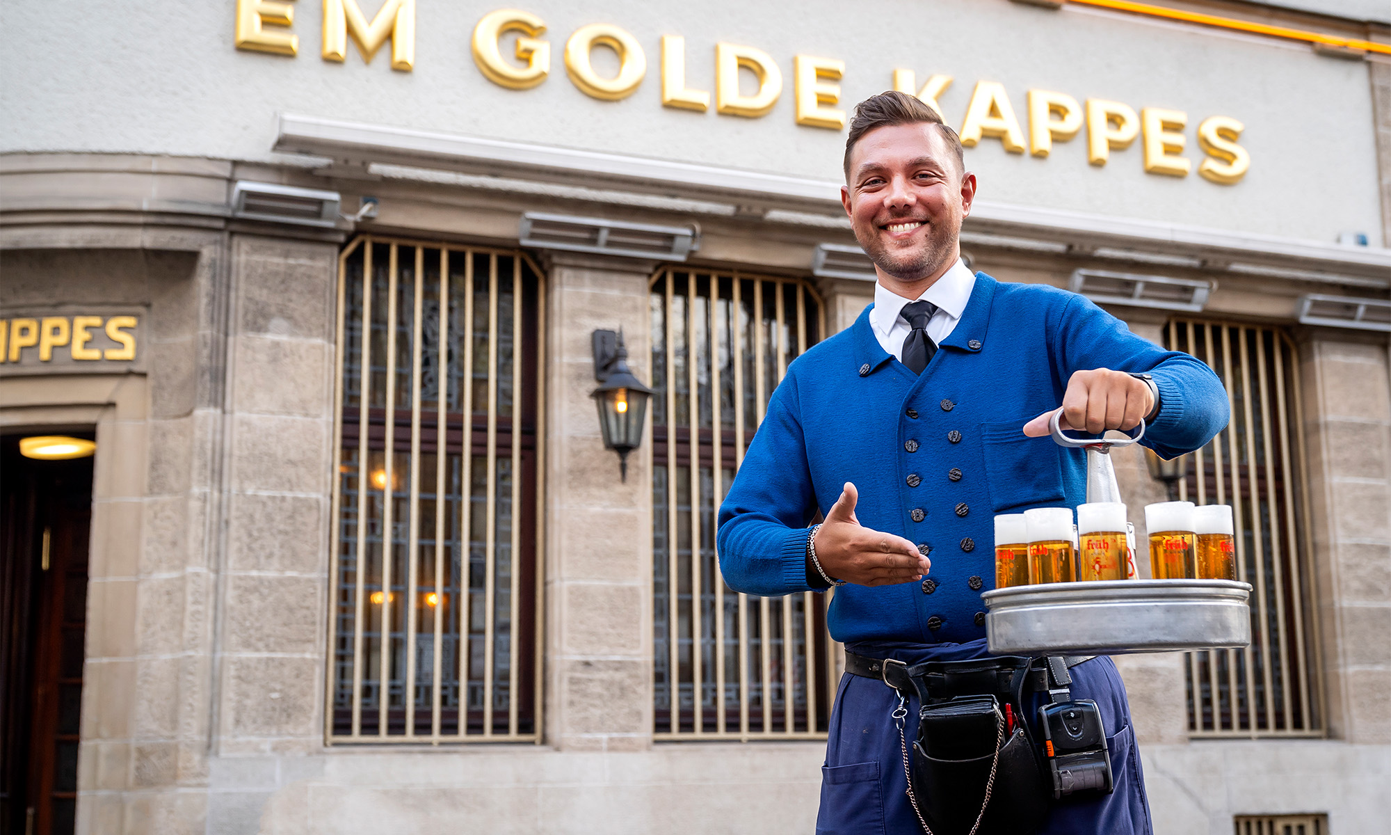 Ein traditioneller Köbes mit einem Kranz voller Kölsch vor dem Gasthaus "Em golde Kappes".