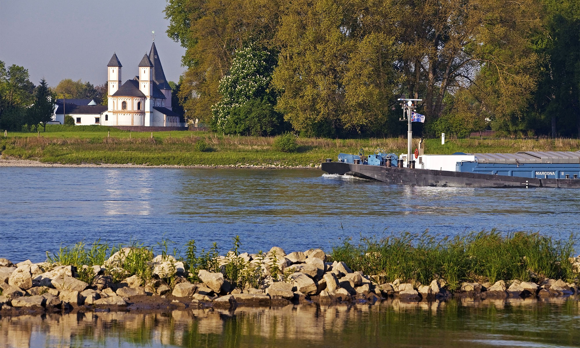 Ein Schiff fährt auf dem Rhein vorbei an der Kirche St. Amandus im Kölner Stadtteil Rheinkassel.