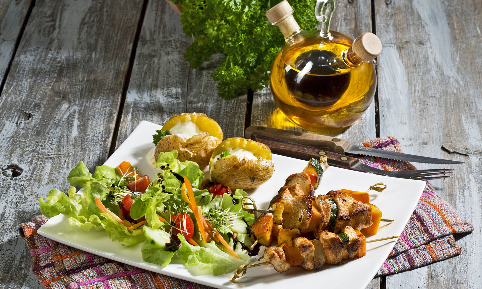 Ein Teller mit Salat, Fleischspieß und Ofenkartoffel, im Hintergund eine Flasche mit Öl.