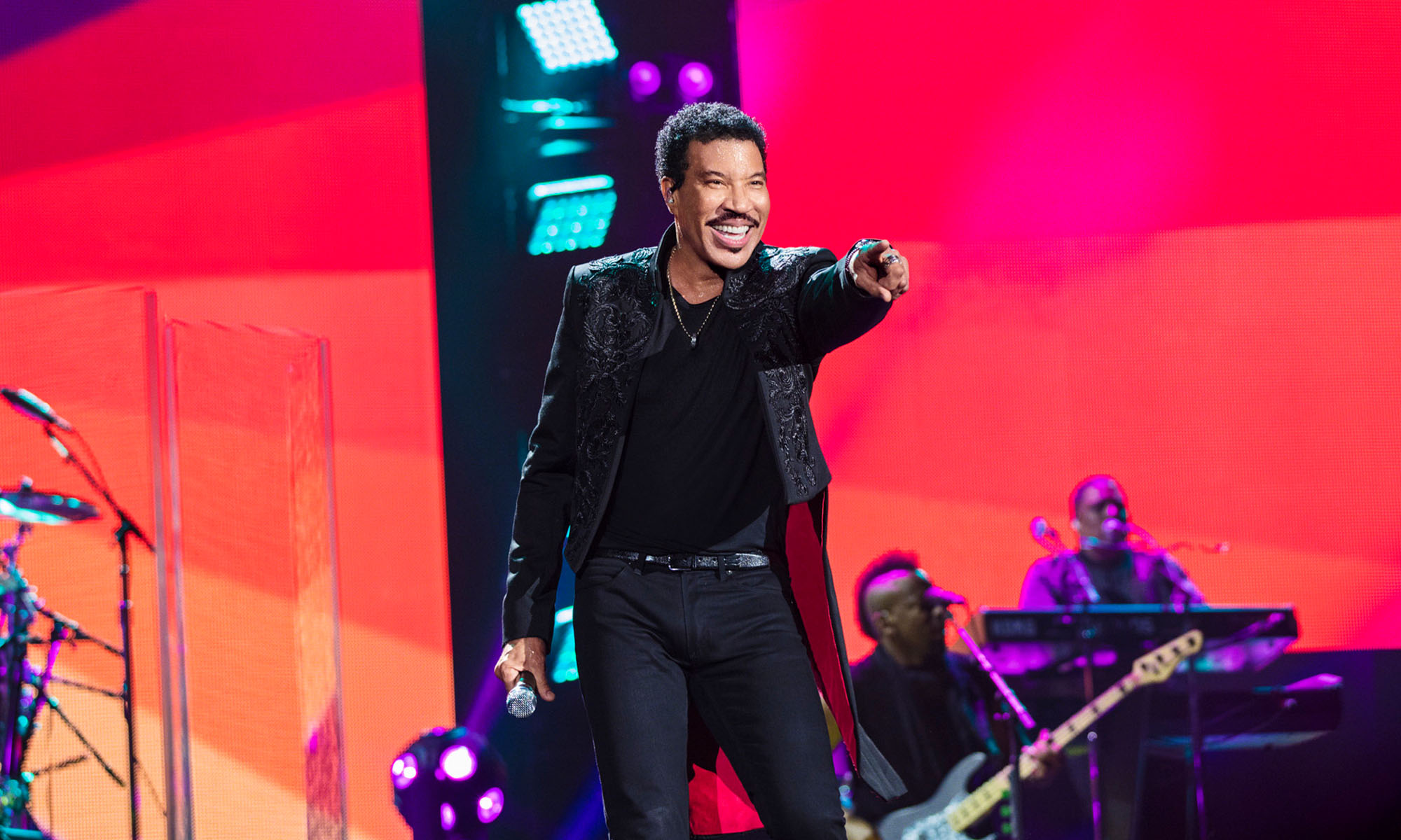 Der Sänger Lionel Richie steht auf der Bühne, die einen roten Hintergrund hat.