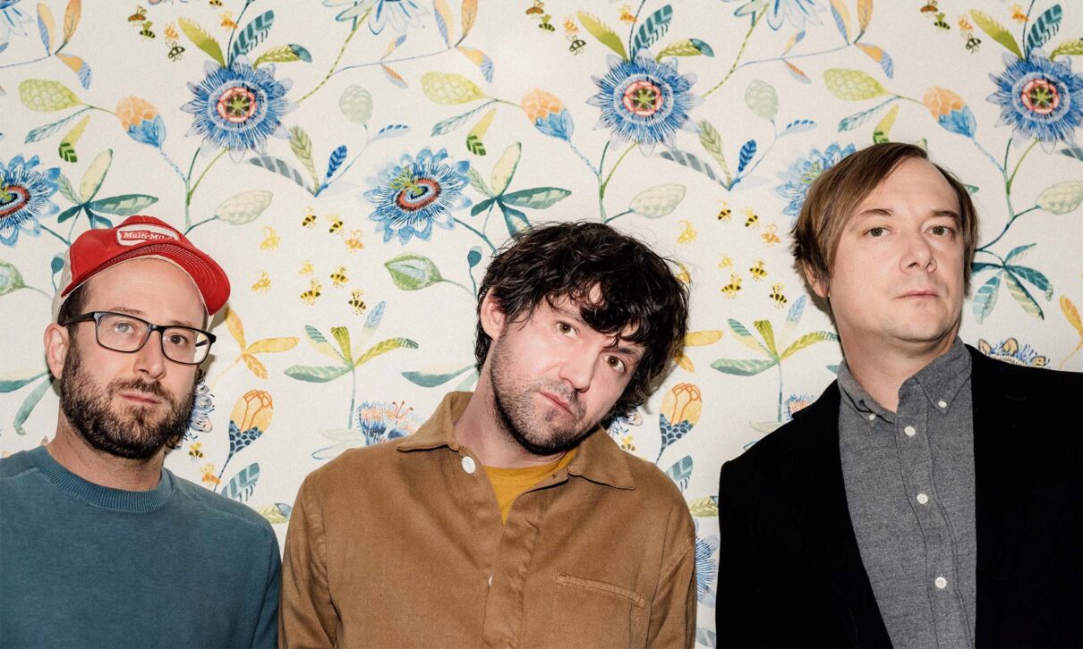 Die drei Mitglieder der US-amerikanischen Band Bright Eyes vor einer bunt gemusterten Tapete.