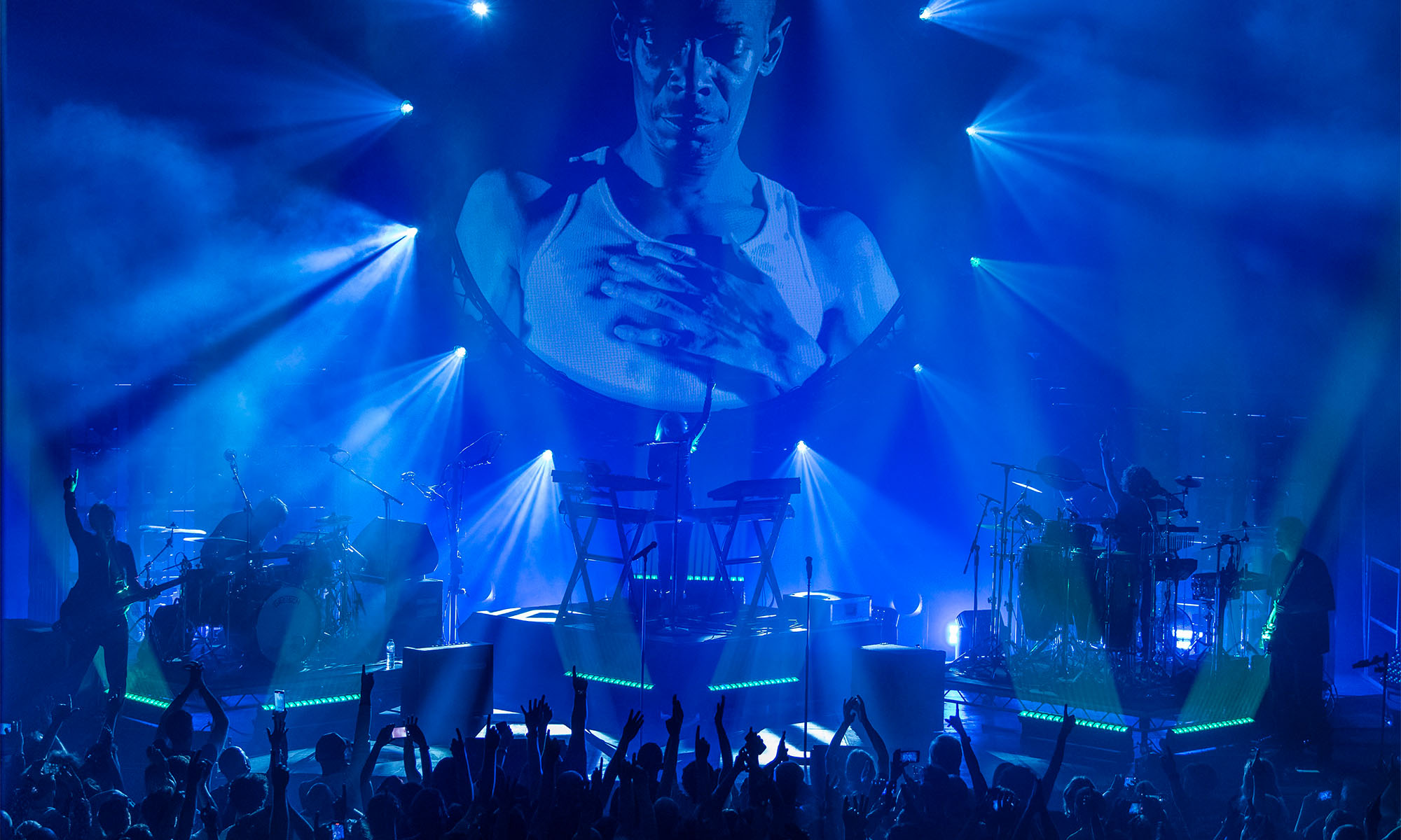 Die Band Faithless gibt ein Konzert, im Hintergrund der blau beleuchteten Bühne ist ein Porträt des verstorbenen Sängers Maxi Jazz zu sehen.
