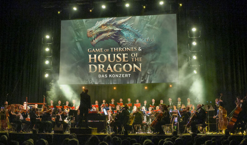 Ein Orchester spielt live die Msuik zu Game of Thrones & House of the Dragon, das auf der Leinwand gezeigt wird.