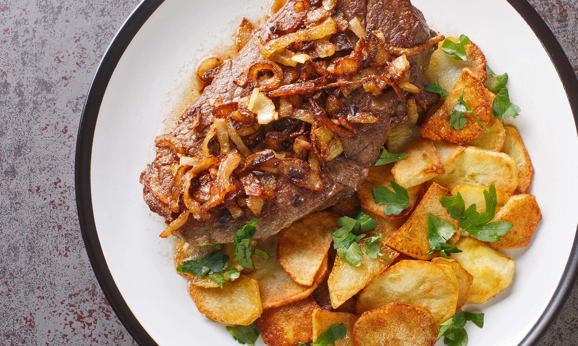 Ein Rostbraten mit Zwiebeln sowie mit Bratkartoffeln als Beilage