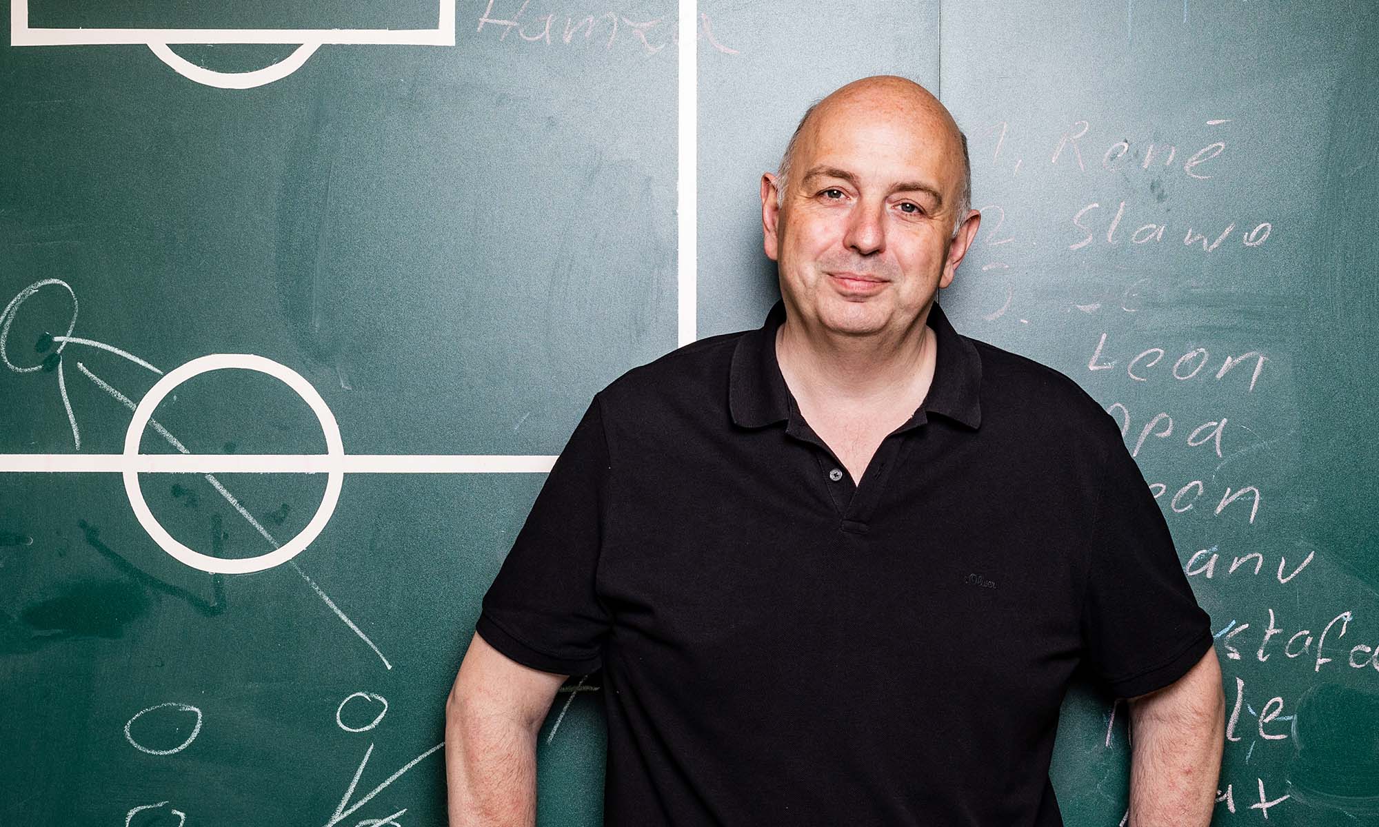 Der Kabarettist Frank Goosen steht vor einer Schultafel, auf der Spielfeldmarkierungen eines Fußballfelsd zu erkennen sind.