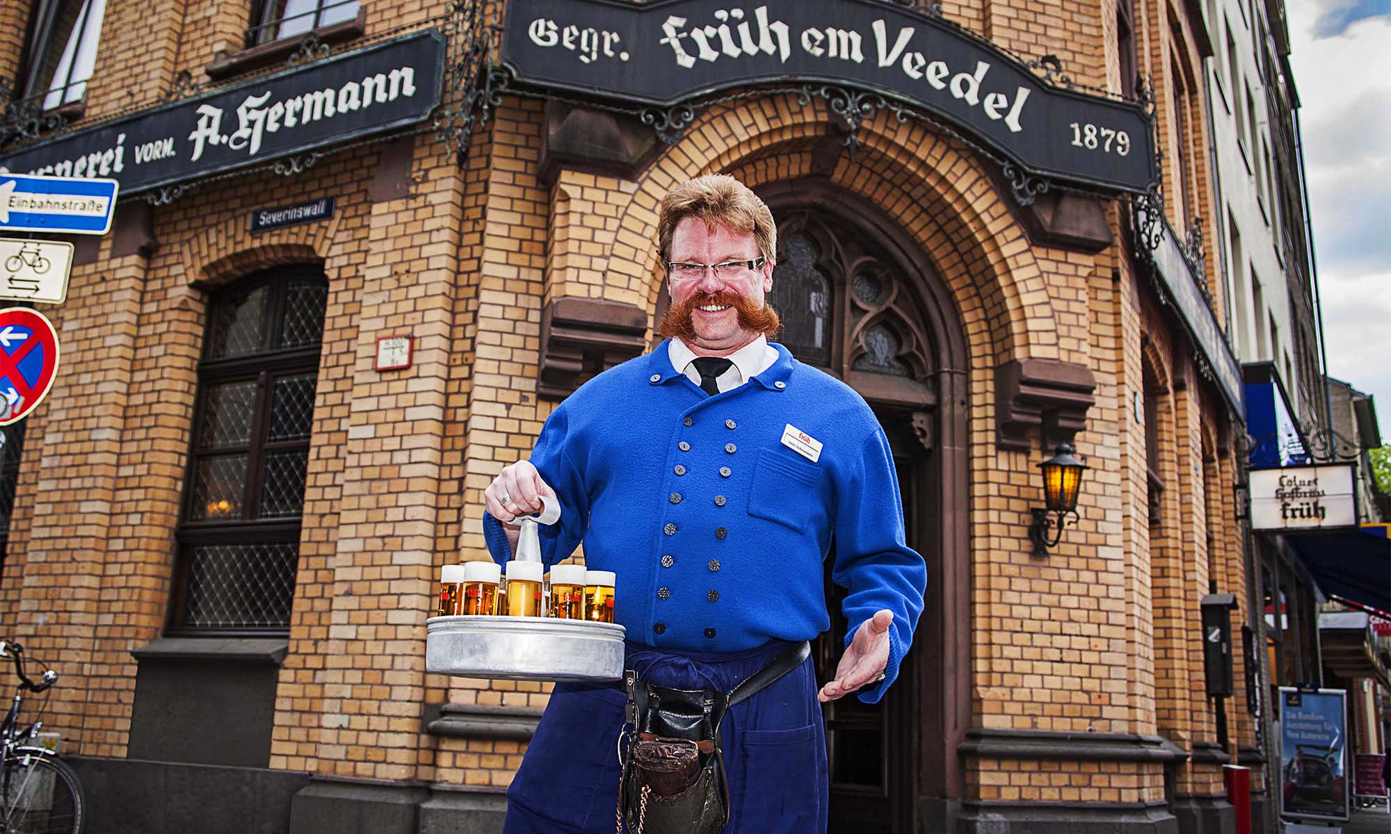 Ein Köbes mit einem Kranz Kölsch steht vor dem Früh em Veedel