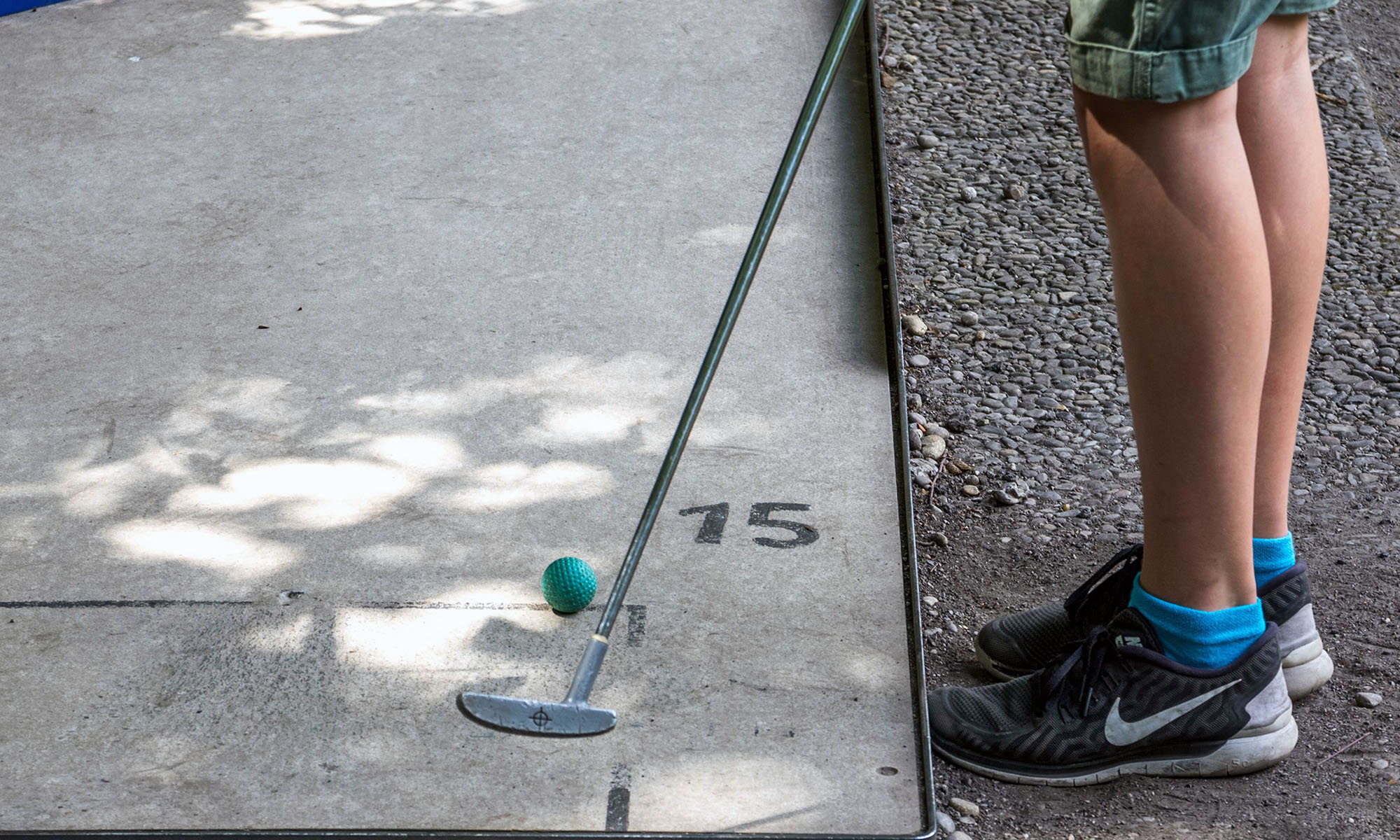 Zu sehen die Beine eines Jungen, der mit einem Minigolfschläger einen grünen Ball spielt