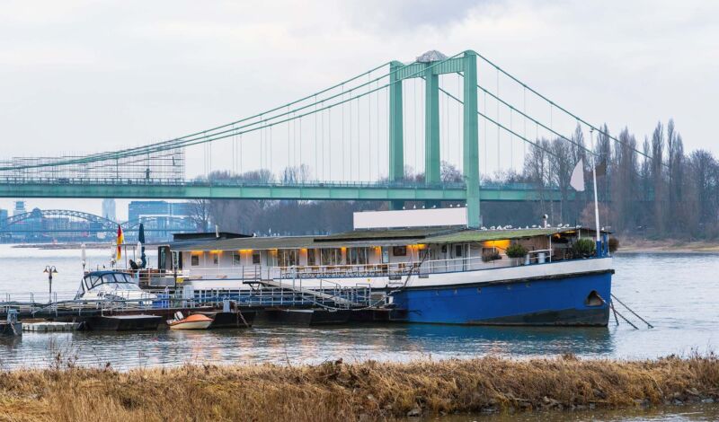 Das Bootshaus MS Rodenkirchen liegt vertäut am Ufer im Kölner Stadtteil Rodenkirchen. Im Hintergrund sieht man die Rodenkirchener Brücke.