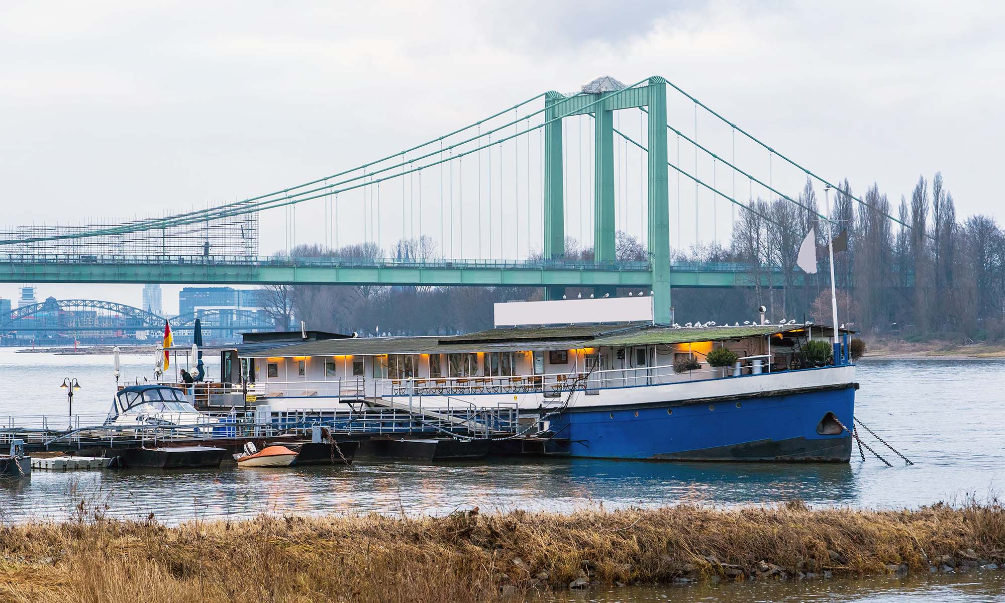 Bootshaus MS Rodenkirchen Das Bootshaus MS Rodenkirchen liegt vertäut am Ufer im Kölner Stadtteil Rodenkirchen. Im Hintergrund sieht man die Rodenkirchener Brücke.