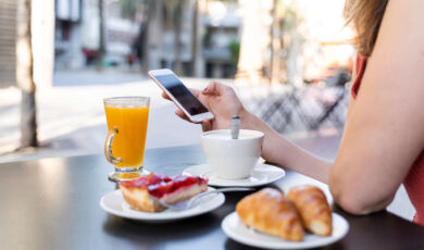 Eine Frau sitzt an einem Tisch mit Kaffeetasse, Orangensaft, Croissant und Kuchen und hält ein Handy in der Hand