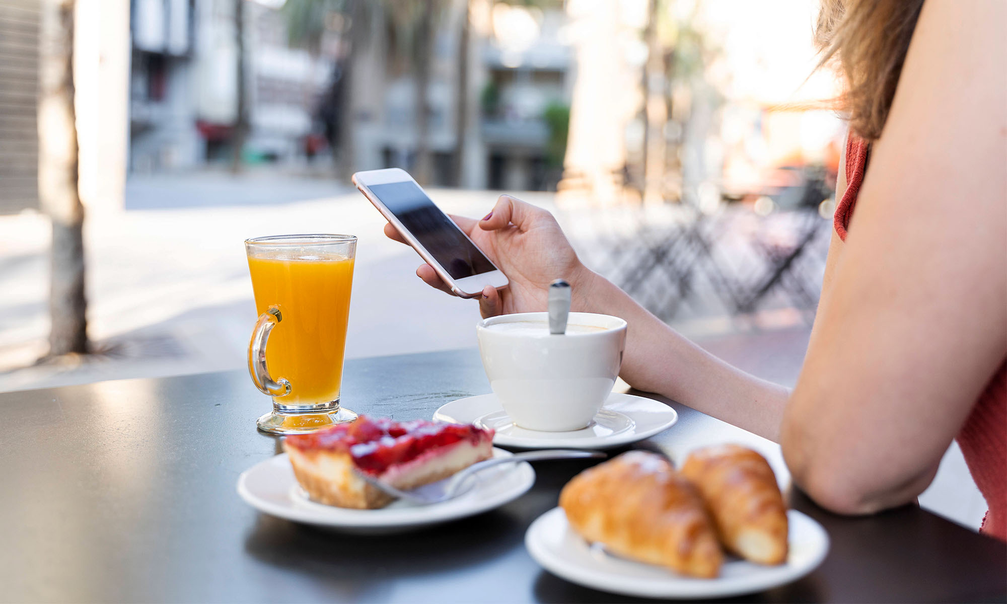 Eine Frau sitzt an einem Tisch mit Kaffeetasse, Orangensaft, Croissant und Kuchen und hält ein Handy in der Hand