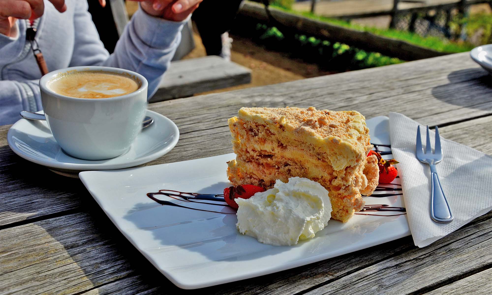 Ein Stück Torte sowie eine Tasse Kaffe auf einem Holztisch, es ist Sommer.