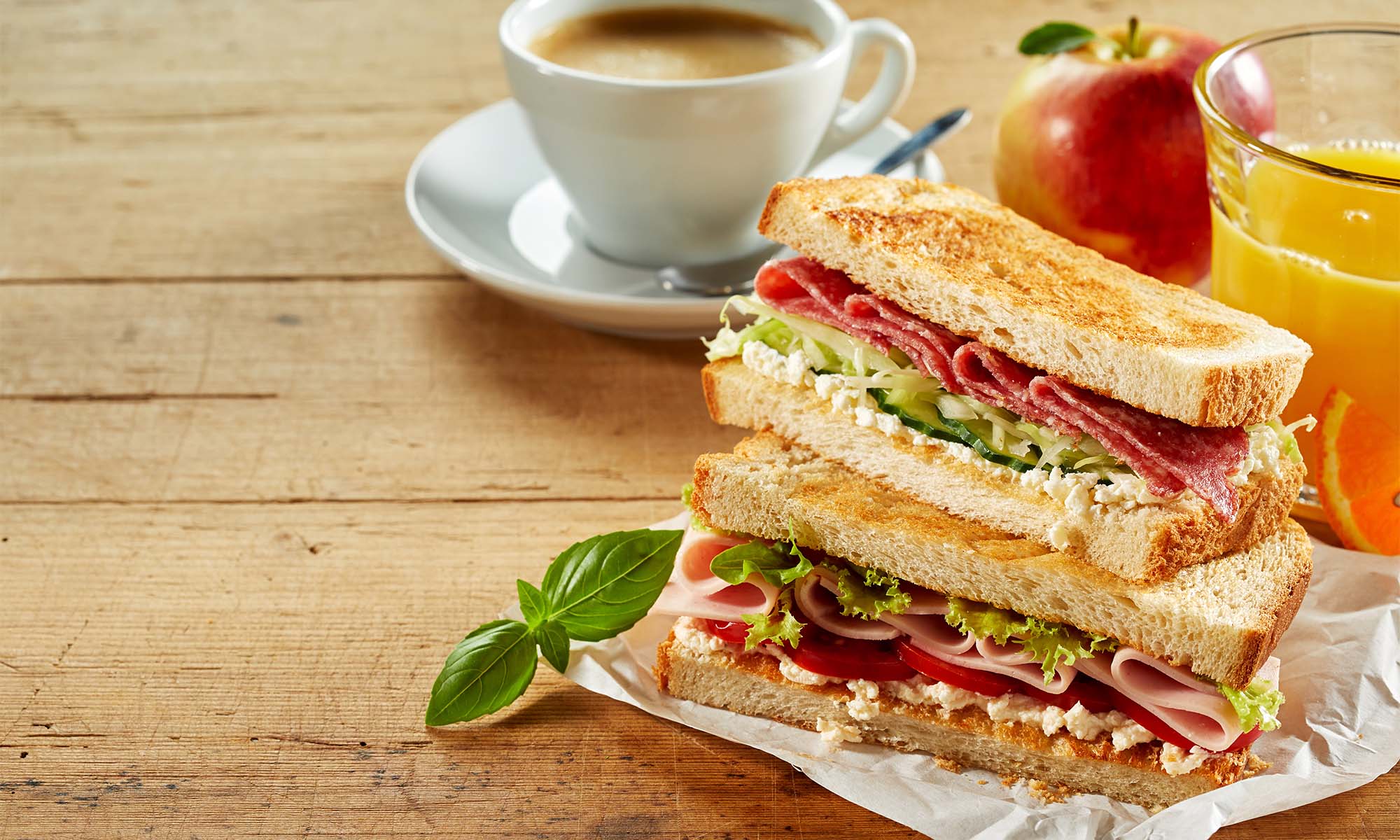 Ein Sandwich mit Wurst, Frischkäse, Gurken und Tomate neben einer Tasse Espresso.