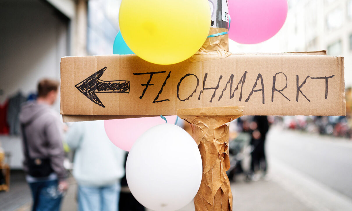Ein handgeschriebens Schild mit dem Text "Flohmarkt" hängt an einem Mast, umgeben von Luftballons.