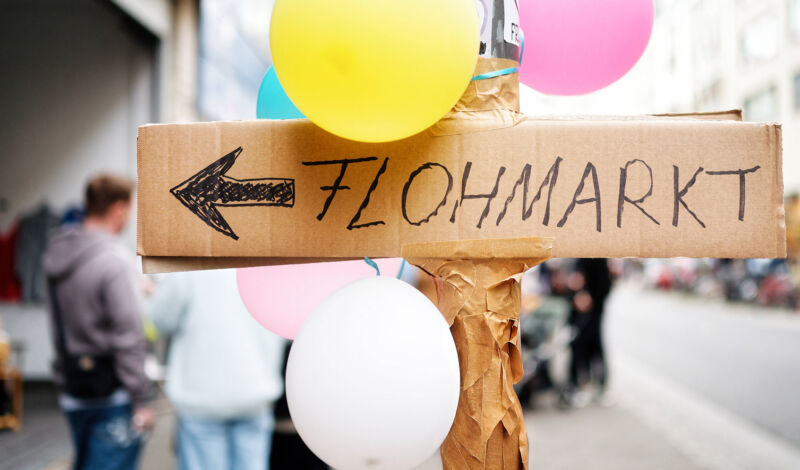 Ein handgeschriebens Schild mit dem Text u0022Flohmarktu0022 hängt an einem Mast, umgeben von Luftballons.