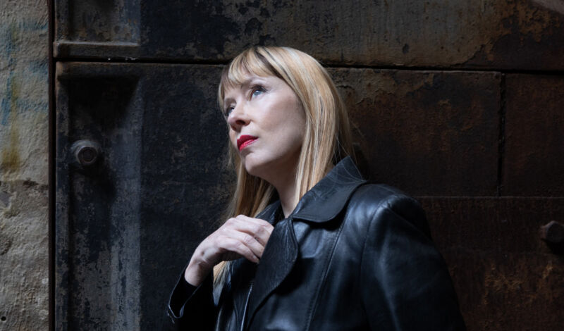 Die amerikanische Sängerin Suzanne Vega in einem schwarzen Mantel vor einem dunklen Hintergrund