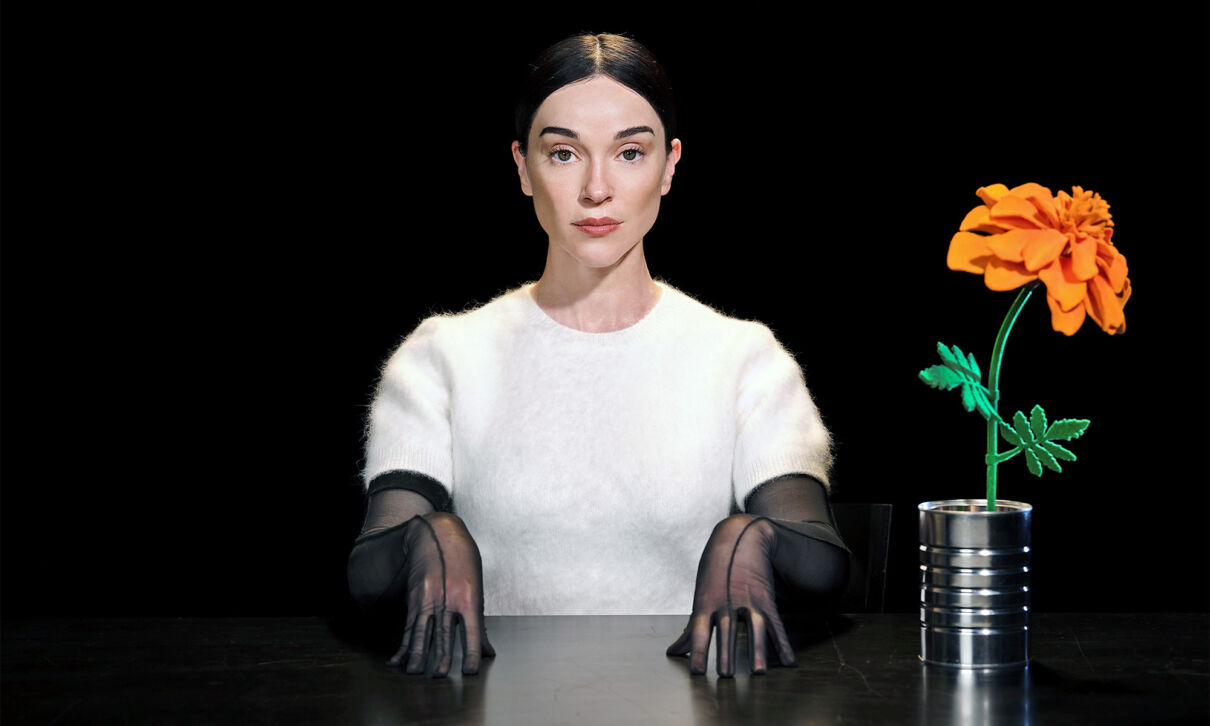 Die Musikerin Annie Clark alias St. Vincent sitzt in einem weißen Pullover mit schwarzen Handschuhen an einem Tisch vor einem schwarzen Hintergrund