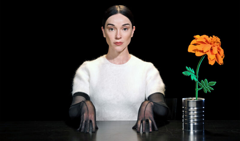 Die Musikerin Annie Clark alias St. Vincent sitzt in einem weißen Pullover mit schwarzen Handschuhen an einem Tisch vor einem schwarzen Hintergrund