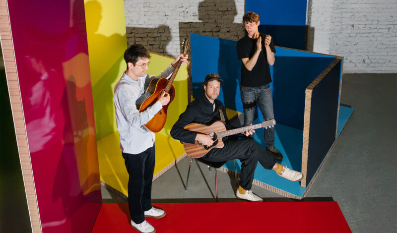 Die drei Musiker der Band AnnenMayKantereit stehen zwischen farbigen Holzwänden in einem Lagerraum.