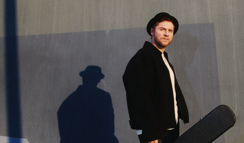 Johannes Oerding mit Gitarrenkoffer vor einer grauen Wand.