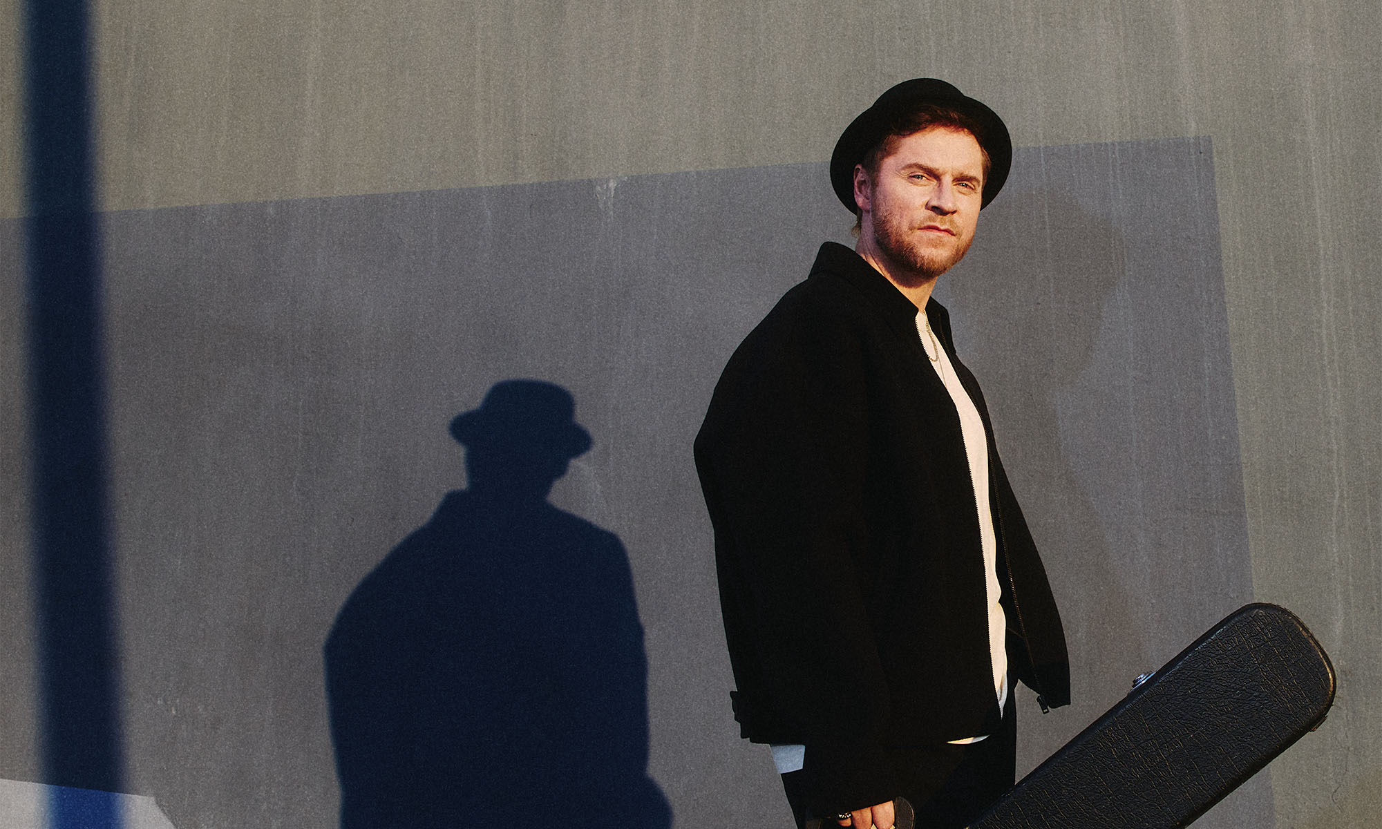 Johannes Oerding mit Gitarrenkoffer vor einer grauen Wand.