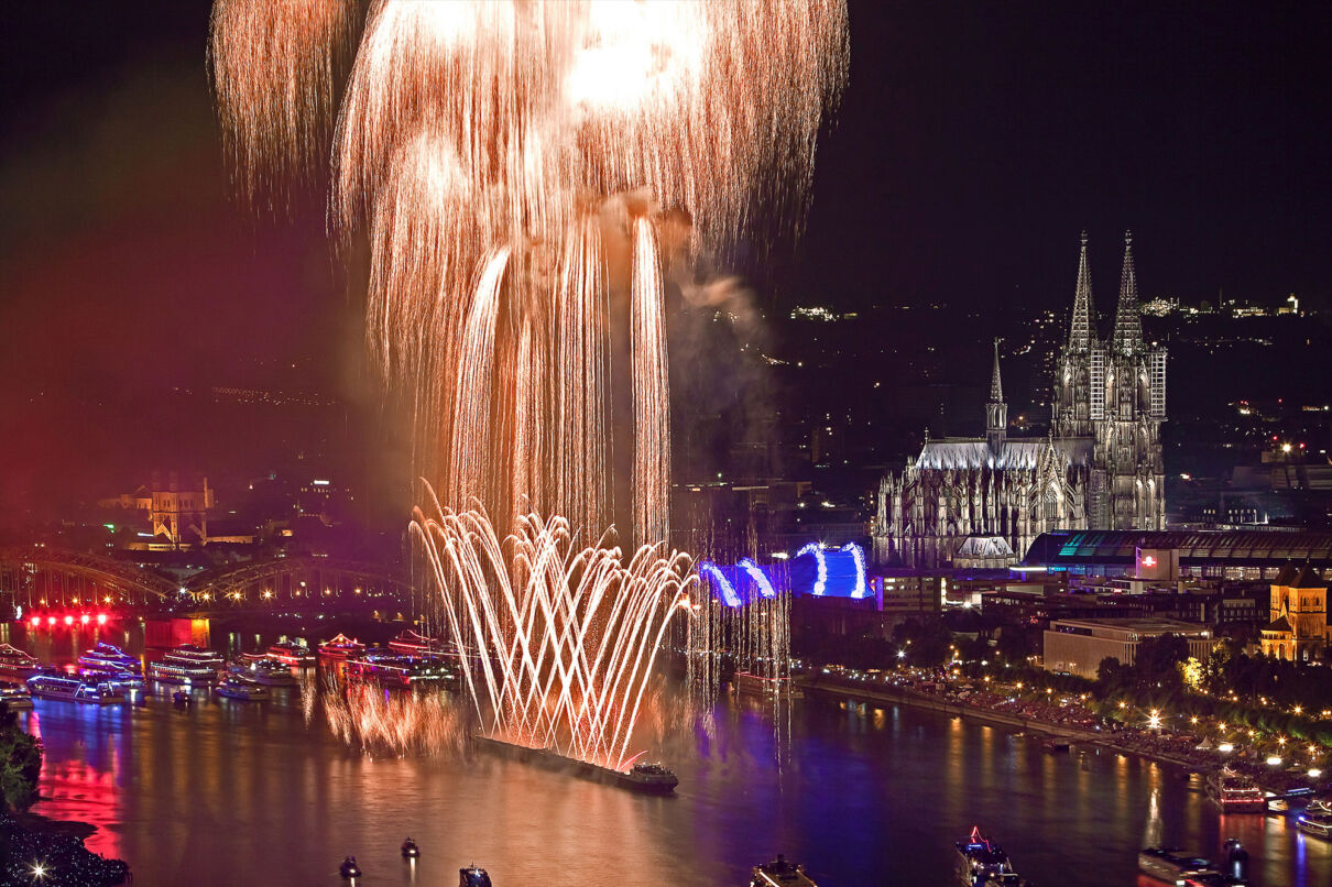 Das Foto zeigt ein Rheinschiff vor der Kulisse der Kölner Altstadt mit Dom, das Feuerwerk in den nächtlichen Himmel schießt.