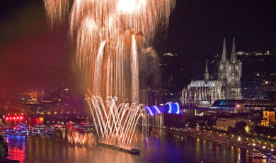 Das Foto zeigt ein Rheinschiff vor der Kulisse der Kölner Altstadt mit Dom, das Feuerwerk in den nächtlichen Himmel schießt.