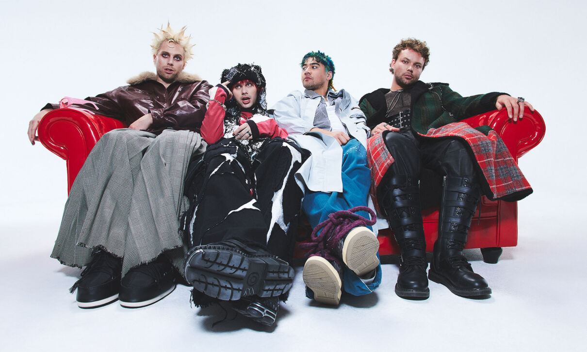 Die Musiker der Band 5 Seconds of Summer sitzen in seltsamer Kleidung auf einem roten Sofa.