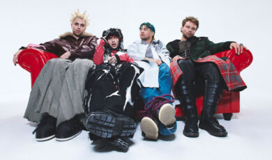 Die Musiker der Band 5 Seconds of Summer sitzen in seltsamer Kleidung auf einem roten Sofa.