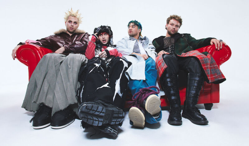 Die Musiker der Band 5 Seconds of Summer sitzen in seltsamer Kleidung auf einem roten Sofa.