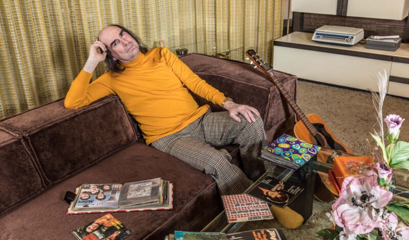 Der Schlagersängersänger Guildo Horn sitzt auf einem Sofa in einem Wohnzimmer, das aussieht wie in den 70er Jahren.