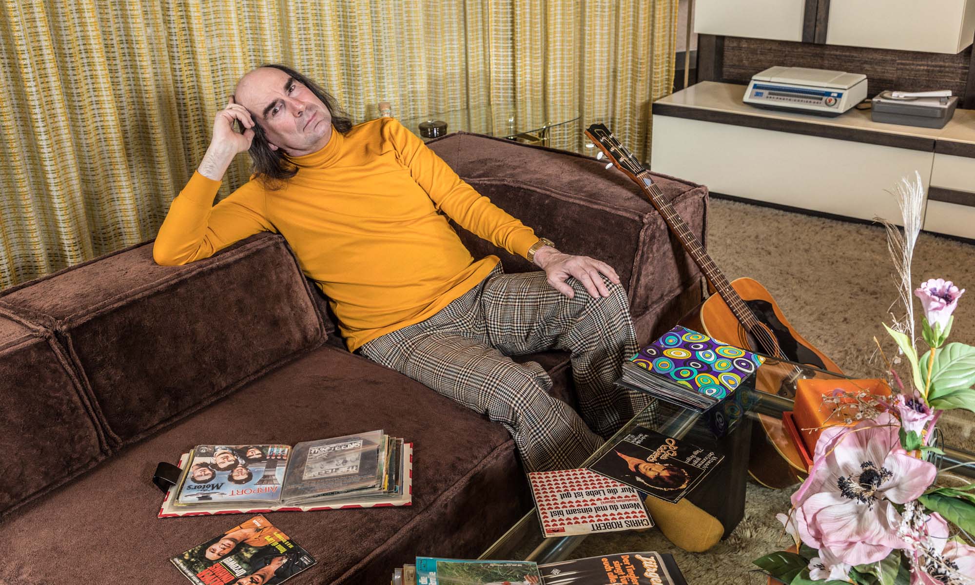 Der Schlagersängersänger Guildo Horn sitzt auf einem Sofa in einem Wohnzimmer, das aussieht wie in den 70er Jahren.