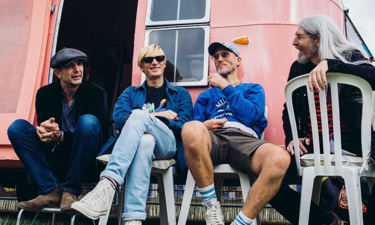 Die Musiker der Band Kula Shaker sitzen auf Plastikstühlen vor einem rosafarbenen Wohnwagen