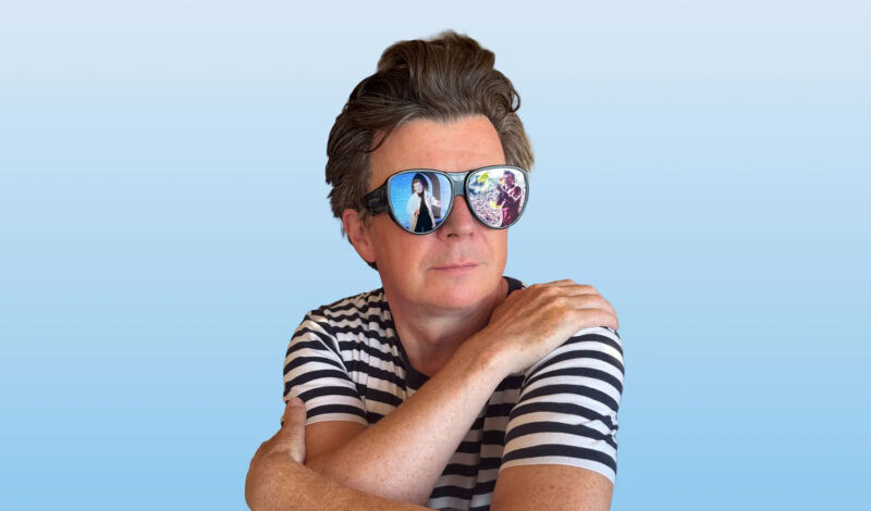 Der britische Popsänger Rick Astlei mit T-Shirt und Sonnenbrille vor blauem Hintergrund.