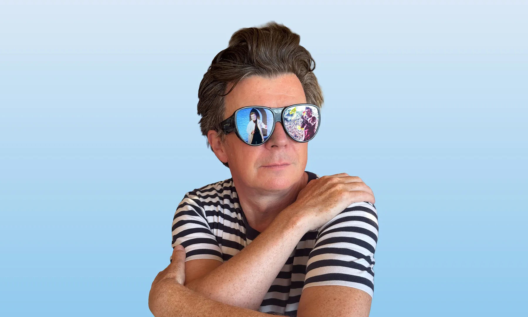 Der britische Popsänger Rick Astlei mit T-Shirt und Sonnenbrille vor blauem Hintergrund.