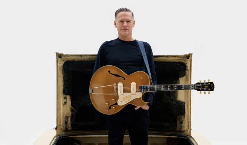 Bryan Adams mit einer Gitarre steht im Kofferaum eines Autos