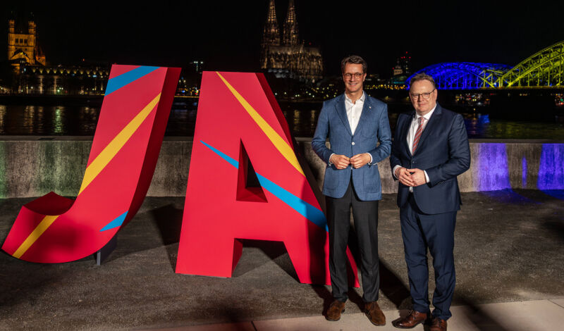 Ministerpräsident Hendrik Wüst und OB Torsten Burmester mit JA Logo vor der illuminierten Hohenzollernbrücke an der Deutzer Rheinpromenade.