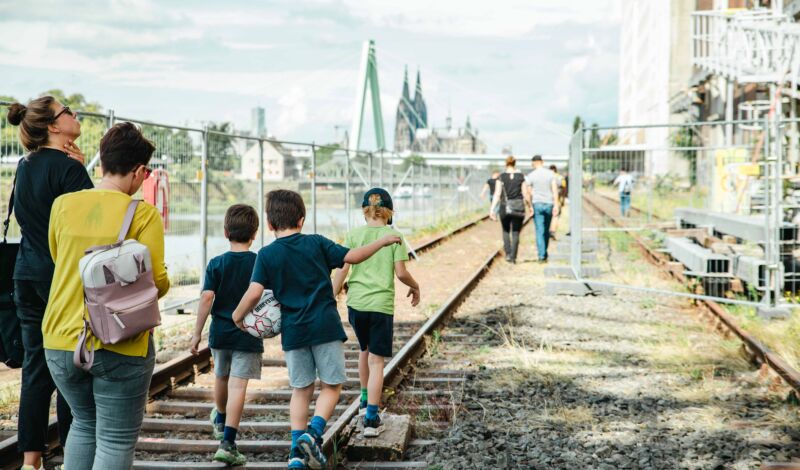 Das Foto zeigt Kinder und Erwachsene, die am Rheinufer entlang des Deutzer Hafens auf Gleisen laufen.