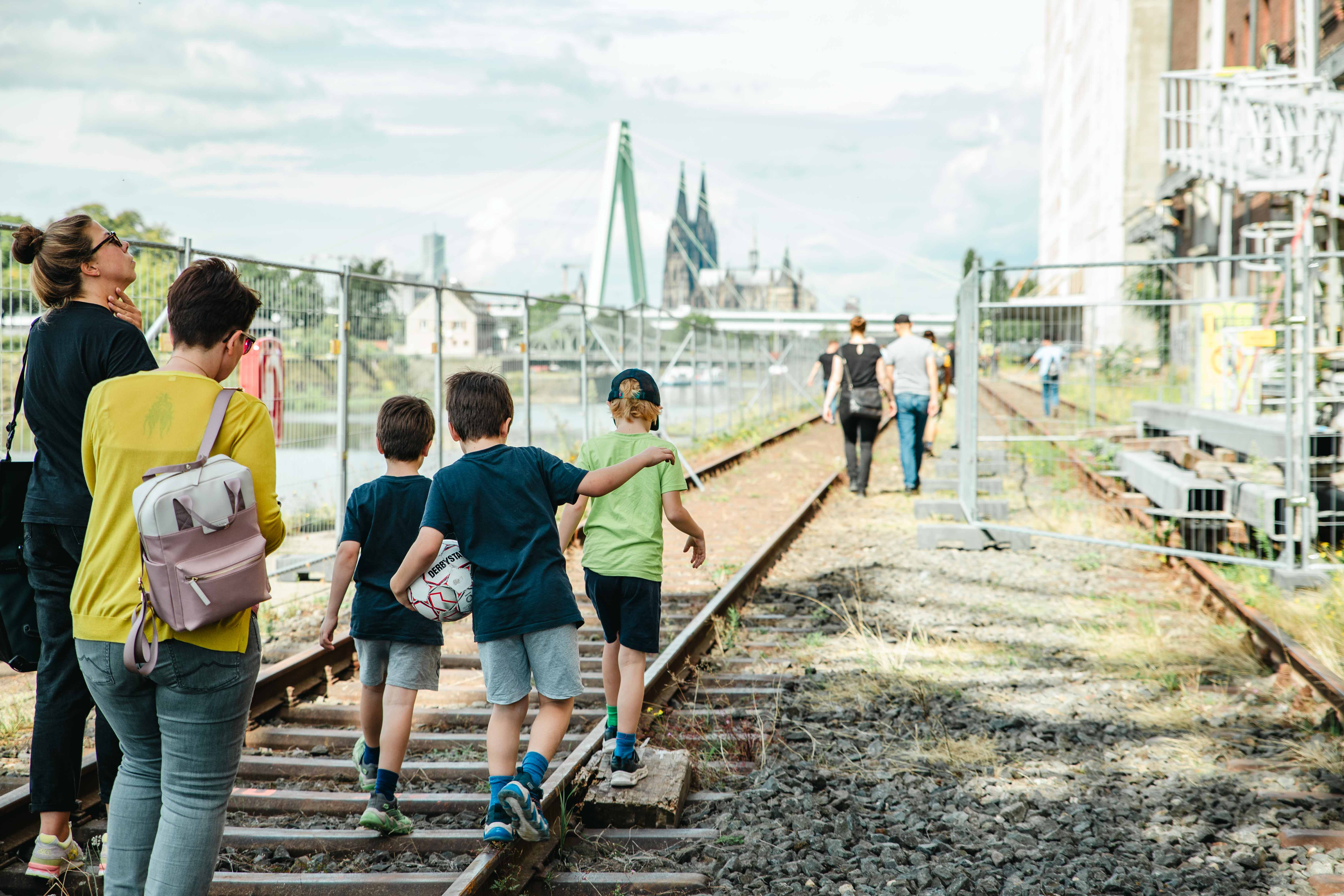 Das Foto zeigt Kinder und Erwachsene, die am Rheinufer entlang des Deutzer Hafens auf Gleisen laufen.